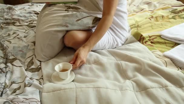 Seductive girl dressed in towel with neat body sitting on a bed, reading magazine and drinking hot tea or coffee - Footage, Video