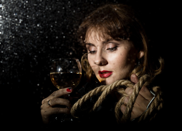 mysterious young woman with a glass of wine posing behind transparent glass covered by water drops. on a dark background - Foto, Bild