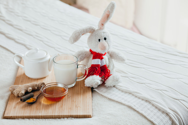 Hot milk in a glass cup and honey on a wooden board. Treatment of children a hot drink. Treatment of folk remedies in bed. Soft toy bunny for your child. - Foto, imagen