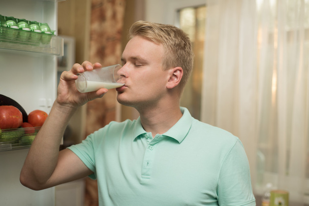 Young man drinking milk in the kitchen, standing at the fridge - Φωτογραφία, εικόνα