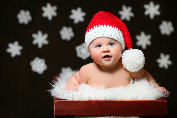 an exited christmas baby sitting in a white wooden box wearing a red tailed hat - Photo, Image