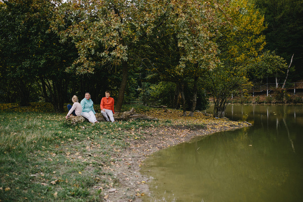 kız, kırmızı kazak giyen genç bir adam ve gölün yanında bir kütükte oturan yaşlı bir kadın. kızı, baba ve büyükanne parkta açık havada eğlenmek  - Fotoğraf, Görsel