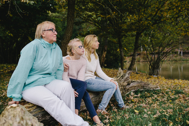 girl, young woman and mature woman sitting on a log in the park. Daughter, mother and grandmother relax outdoor on the background of autumn forest and lake and look forward - Foto, Imagem