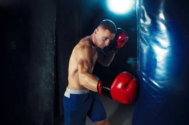 Male boxer boxing in punching bag with dramatic edgy lighting in a dark studio - Фото, изображение
