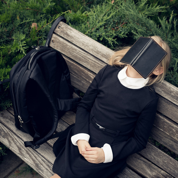 schönes blondes Mädchen in Schuluniform sitzt auf Parkbank und versteckt sich hinter Buch . - Foto, Bild