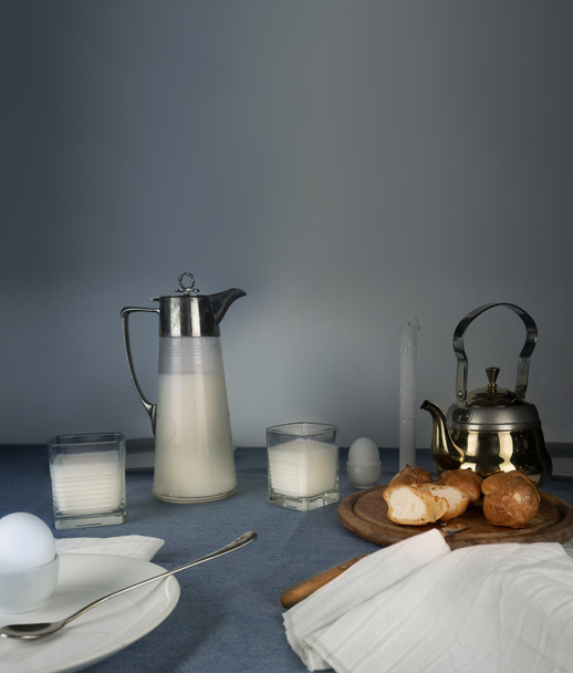 still life. rustic dinner. milk jug, candles, tea, eggs, bread rolls on the table - Foto, imagen
