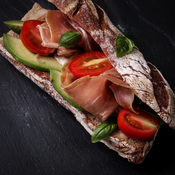 Ciabatta sandwich with prosciutto,avocado,cherry tomatoes and basil on dark background - Fotoğraf, Görsel