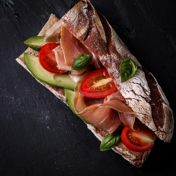 Ciabatta sandwich with prosciutto,avocado,cherry tomatoes and basil on dark background - Fotoğraf, Görsel