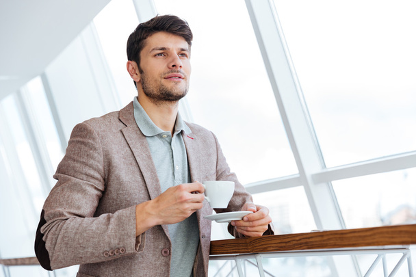 Pensive young businessman in jacket holding coffee cup indoors - Фото, изображение