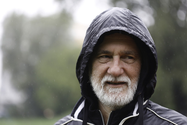 Retrato de un hombre barbudo blanco mayor con una sudadera con capucha negra en un día lluvioso con un fondo borroso
 - Foto, Imagen