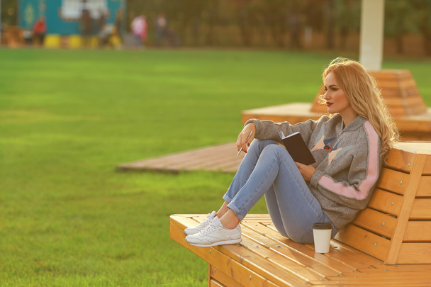Mujer de moda en sentarse en el banco en el parque de la ciudad. Beber café para llevar y escribir o dibujar algo en un cuaderno
. - Foto, Imagen
