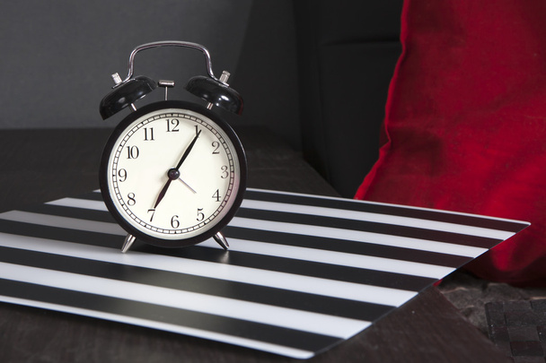 Μαύρο ρολόι συναγερμών σε ένα μαύρο και άσπρο ριγέ χαρτοπετσέτα δείχνει 7 η ώρα στο κομοδίνο - Φωτογραφία, εικόνα
