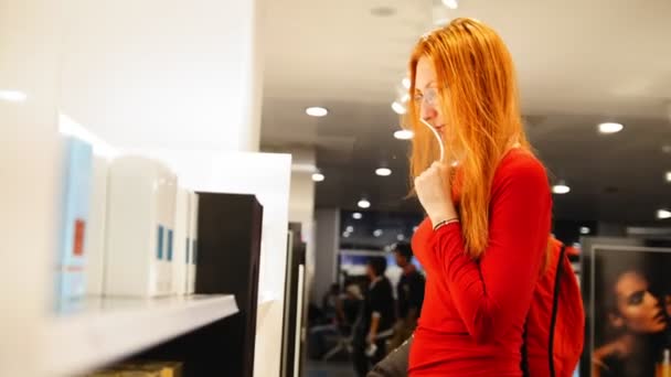 Beautiful red hair girl in glasses choosing a perfume in duty free area at airport - Πλάνα, βίντεο