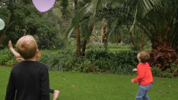 Three young children having fun hitting balloons in a park - slomo - Кадры, видео