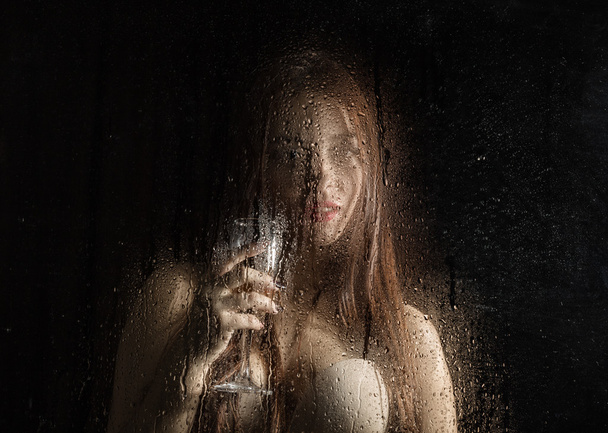 Smooth portrait of sexy model, posing behind transparent glass covered by water drops. young woman holding a glass of champagne - Photo, Image