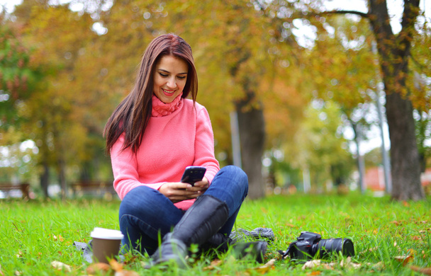 Young fashionable teenage girl with smartphone, camera and takeaway coffee in park in autumn sitting at smiling. Woman in fall in park texting - Photo, Image