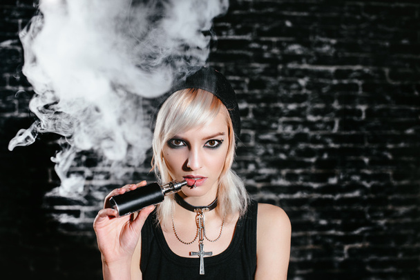 Sexy goth girl smokes electronic cigarette on dark background. The model vaper vaping a vaporizer in the studio. - Photo, Image