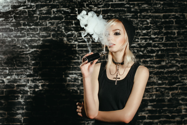 Sexy goth girl fume cigarette électronique sur fond sombre. Le mannequin vaporise un vaporisateur en studio
. - Photo, image