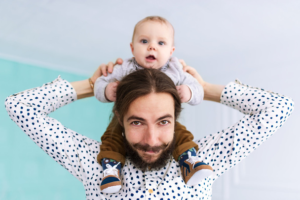 Charming baby son sitting on father neck in white room - Foto, Bild