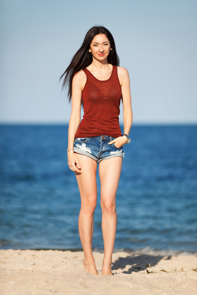 Νέοι όμορφη μόδας γυναίκα να στέκεται στην παραλία. Καλό καλοκαίρι υπαίθρια τρόπο ζωής. Διακοπές. Ωκεάνια παραλία χαλαρώστε. Σέξι μοντέρνα γυναίκα με μακριά μαλλιά σε σορτς jean και κόκκινο τιραντακι που περπατούν στην παραλία - Φωτογραφία, εικόνα