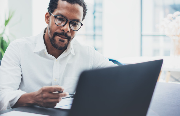 Dark skinned coworker wearing glasses and using laptop in modern office.African american man in white shirt working on workplace.Horizontal,blurred background - Fotoğraf, Görsel