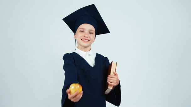 Close up portrait graduate girl 7-8 years in the mantle and hat holding book and gives you apple on white and smiling at camera - Záběry, video