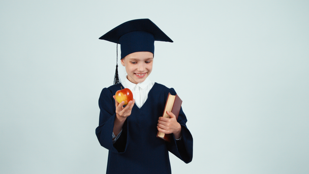 Портрет выпускницы девушка 7-8 лет в мантии и шляпе держа книги и дает вам яблоко на белом и улыбаясь в камеру
 - Кадры, видео