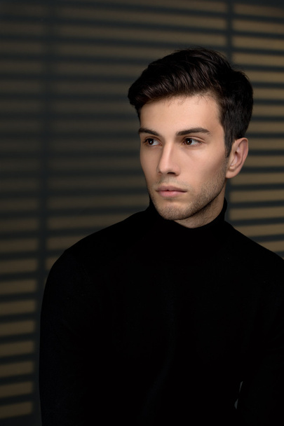 Стильный молодой человек в черном свитере сидит рядом с жалюзи и thinki
 - Фото, изображение