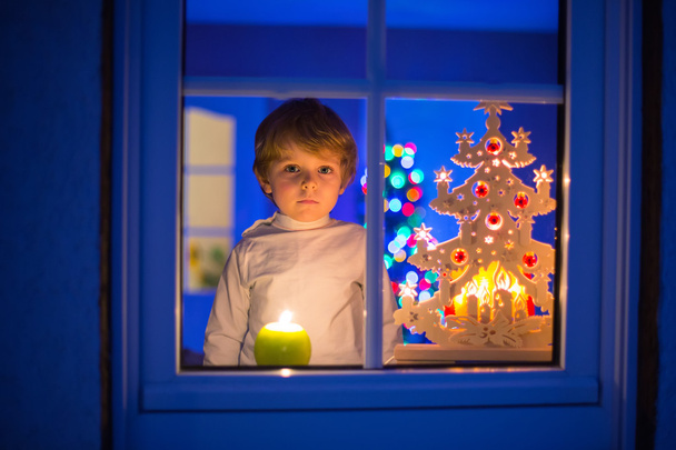 kleiner Junge steht zur Weihnachtszeit am Fenster und hält  - Foto, Bild