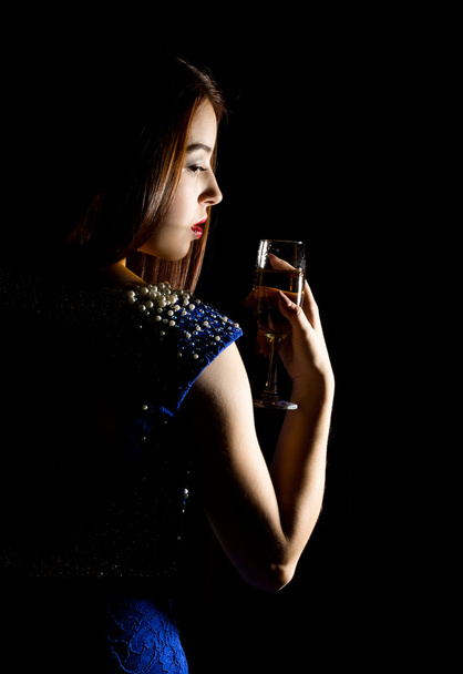 Young celebrating woman in a blue dress holding a glass of champagne on a dark background. play of light and shedow - 写真・画像