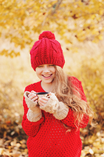 happy child girl in warm red knitted hat and scarf on cozy autumn walk drinking hot tea from thermos. Seasonal outdoor activities - Φωτογραφία, εικόνα