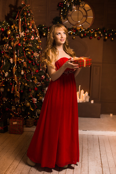 Young beautiful blond woman in the red dress on christmas, woman in a beautiful room with a Christmas tree and candles, a girl with gifts,  golden hair, happy woman new year, Redhead girl smiling  - Photo, image