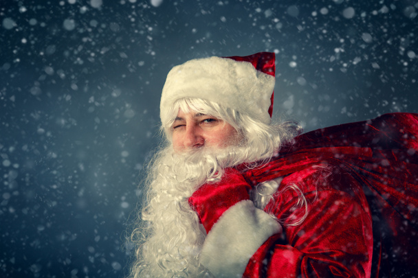 Подмигивающий Санта Клаус носит сумку с подарками
.  - Фото, изображение