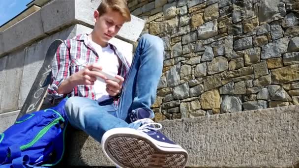 Adolescente menino com skate e mochila sms escrita, mensagens de texto no smartphone sentado em escadas
 - Filmagem, Vídeo