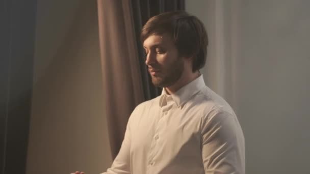 Brutal handsome man dressed in wedding day. The groom goes to the window, she puts on a red tie on a close-up of neck. The camera moves to the left. The frame is filled with light. The Big Day. - Кадри, відео