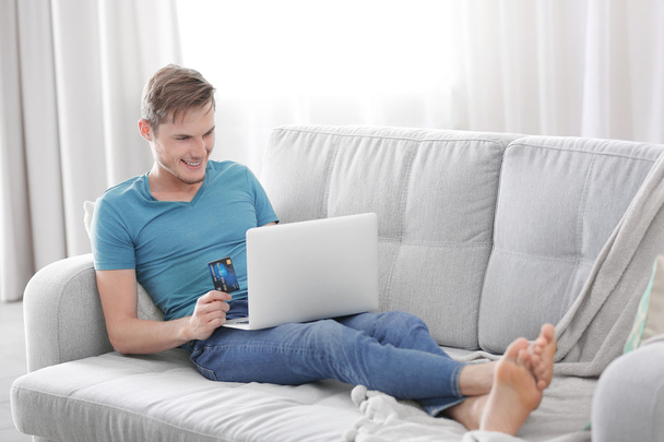 Young man with laptop sitting on sofa and making online shopping - Foto, imagen