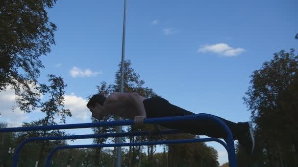 Athletic man doing gymnastics elements on bar in city park. Male sportsman performs strength exercises during workout outdoor. Young guy demonstrates static exercise. Training outside. Slow motion - Footage, Video