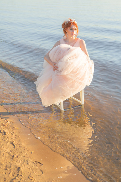 bride sitting on a chair in the water realized dress and watch the sunset - Foto, Imagen