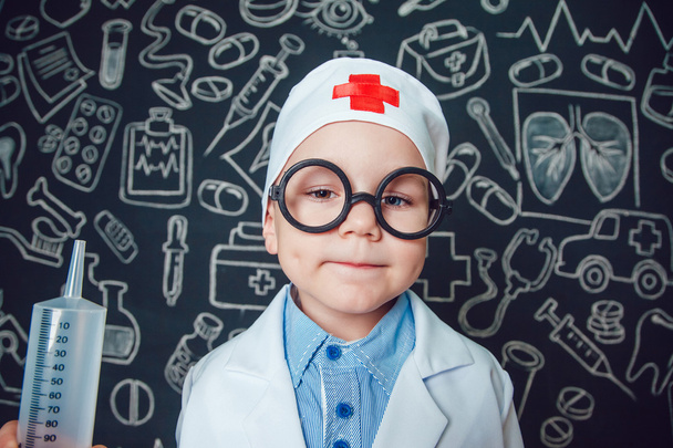 Happy little boy as a doctor in glasses holding syringe on dark background with pattern - Photo, Image