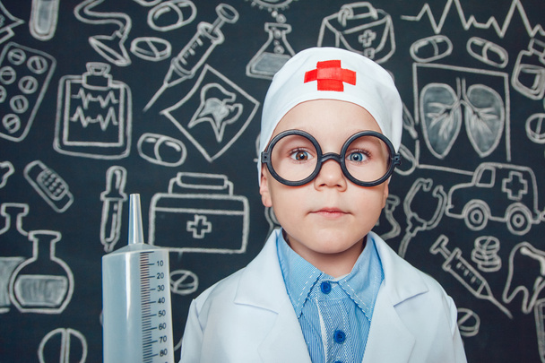 Happy little boy as a doctor in glasses holding syringe on dark background with pattern - 写真・画像