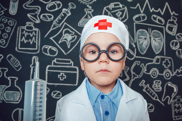 Happy little boy as a doctor in glasses holding syringe on dark background with pattern - Foto, Imagem