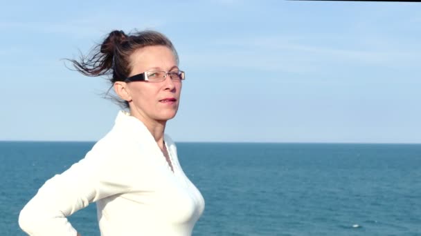 4k. Ενήλικη γυναίκα στα γυαλιά και λευκή μπλούζα ενάντια την επιφάνεια της θάλασσας - Πλάνα, βίντεο