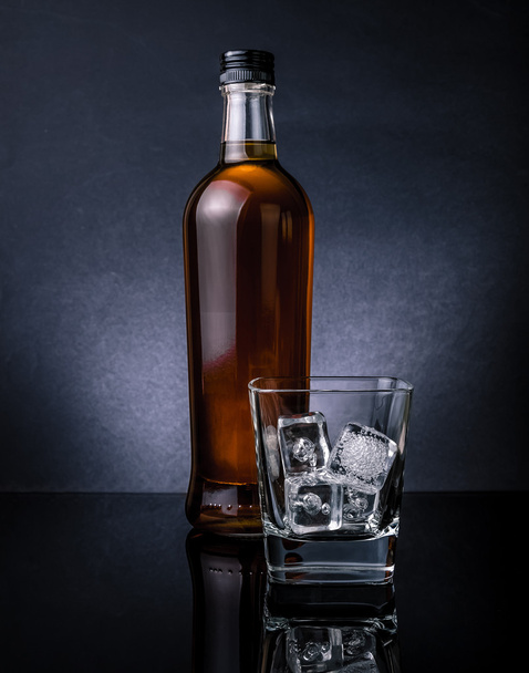 whiskey with ice cubes in glass near bottle on black background, cold atmosphere - Фото, зображення
