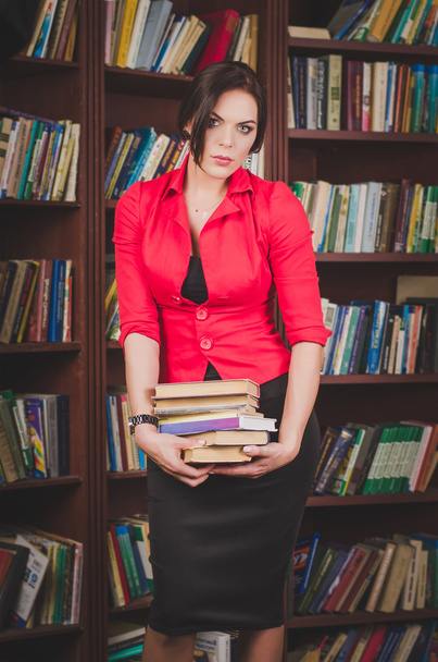 beautiful young brown-haired woman in office attire standing near bookshelves and holds a stack of books - Foto, afbeelding