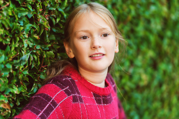 Retrato de cerca al aire libre de la linda niña de 8-9 años usando jersey rojo brillante, de pie junto a arbustos verdes
 - Foto, imagen