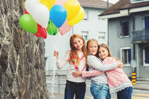 Außenporträt von 3 niedlichen kleinen Mädchen mit bunten Luftballons, die Sweatshirts tragen - Foto, Bild