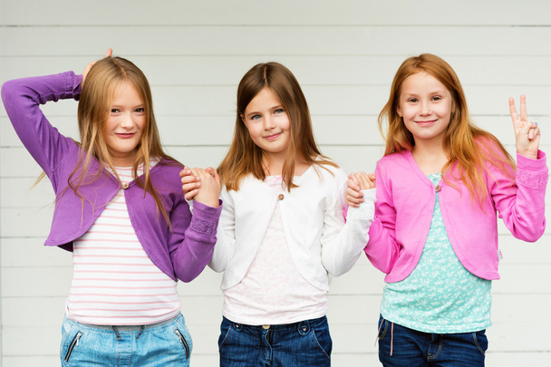Група з трьох маленьких дівчат, що стоять на відкритому повітрі на білому дерев'яному фоні, з джинсовими джинсами та різнокольоровими куртками
 - Фото, зображення