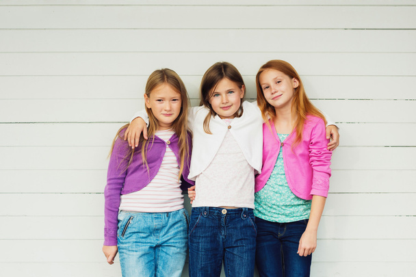Grupo de 3 meninas em pé ao ar livre contra fundo de madeira branca, vestindo jeans jeans jeans e casacos coloridos
 - Foto, Imagem