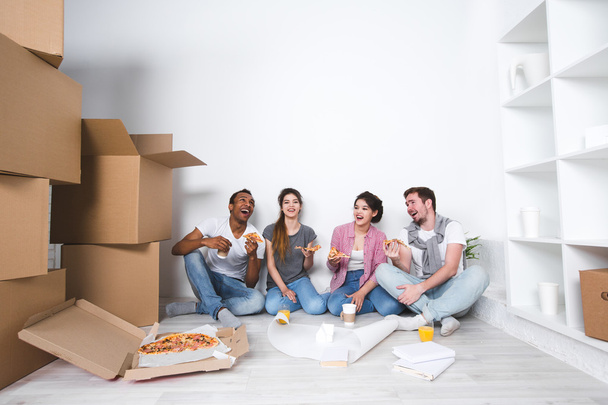 New home. Friends sitting on the floor in the new apartment and eating pizza after unpacking. - Photo, Image