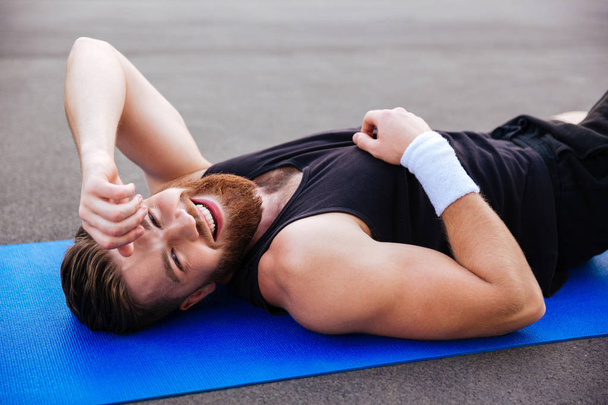 Cheerful laughing sports man resting on blue fitness mat outdoors - Foto, Bild